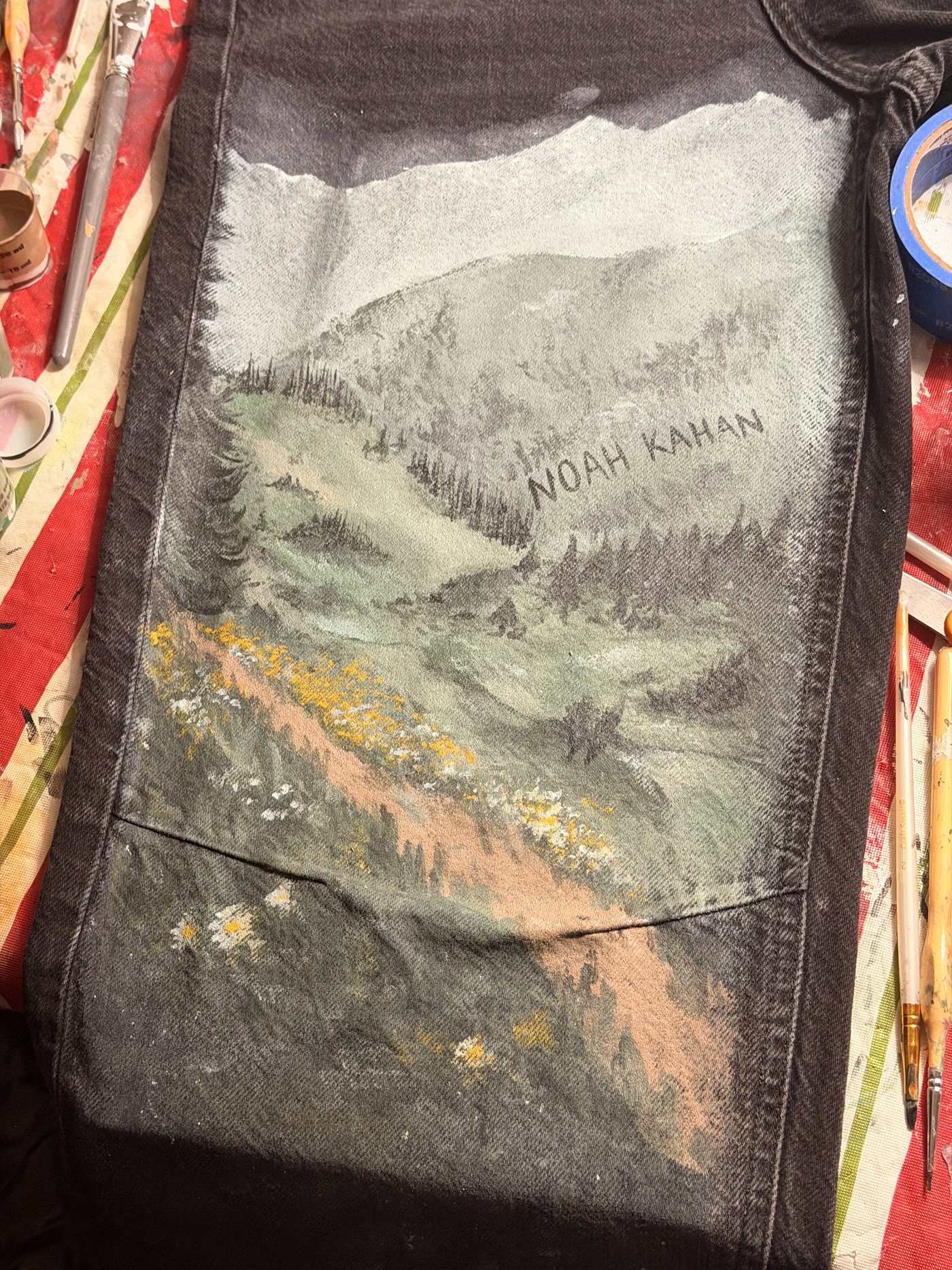 Black jeans hand-painted with a misty mountain landscape, wildflowers, and the name Noah Kahan