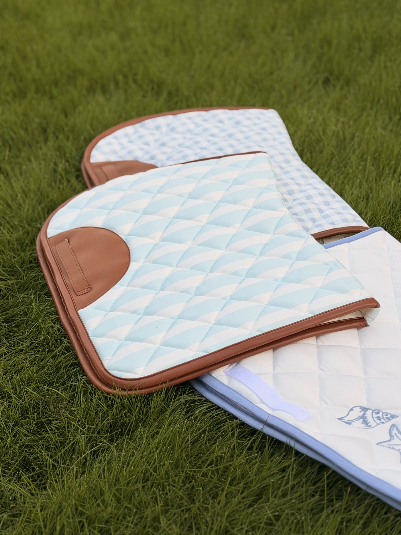 Three Low Tide Equestrian saddle pads laid on grass: striped, gingham, and seashell designs